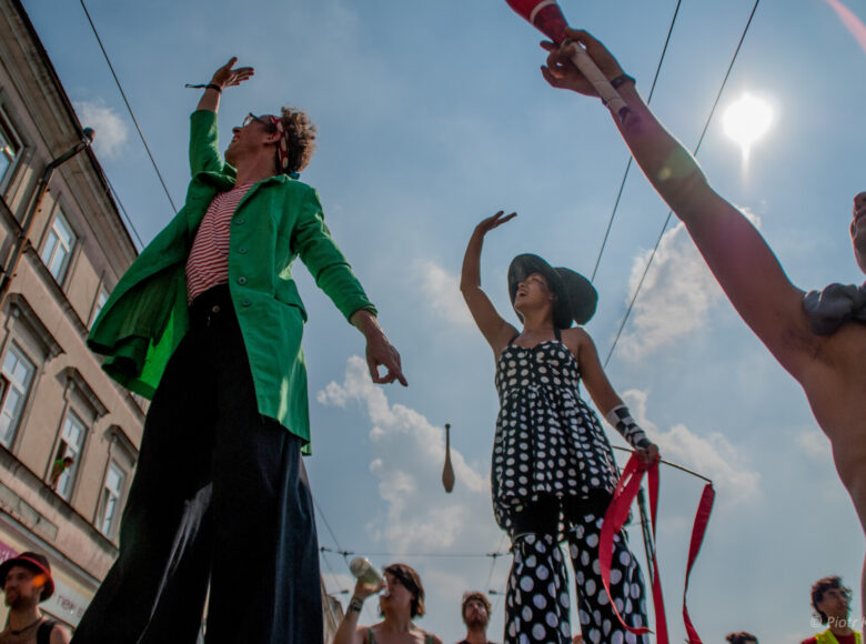 Letni dzień. Pokazani od dołu dziewczyna i chłopak idący na szczudłach oraz chłopak z maczugą żonglerską w ręku. Wszyscy troje machają do okien widocznego obok budynku.