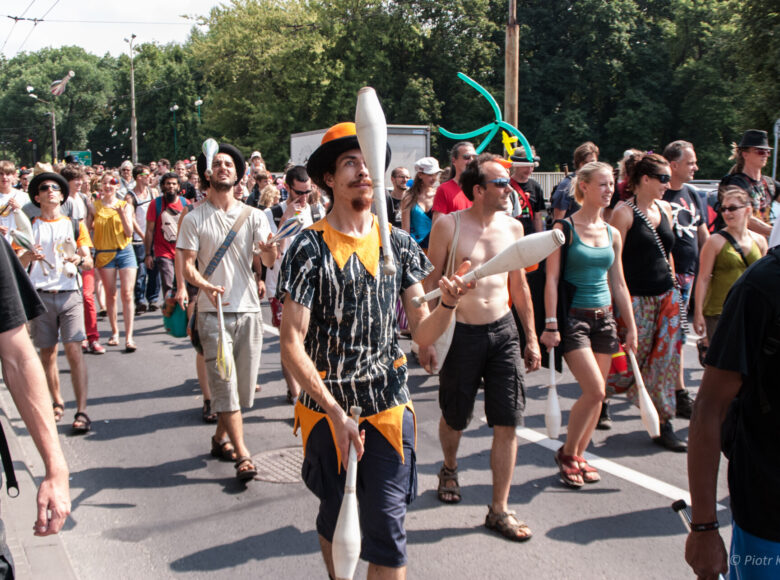 Letni dzień. Ulicą idzie korowód ludzi, wśród nich kilku żonglerów w czarnych kapeluszach. W tle szpaler drzew.