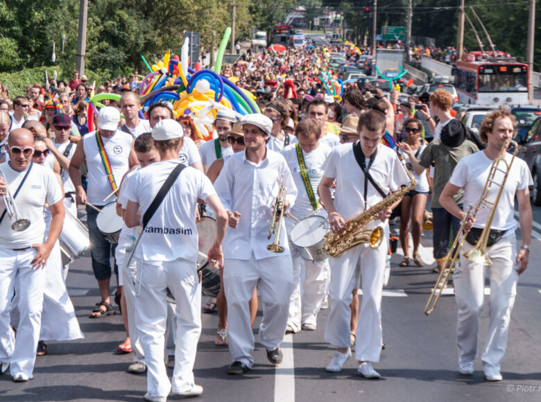 Letni dzień. Ulicą idzie parada, na jej czele grupa ubranych na biało muzyków z instrumentami. Jeden z nich odwrócony jest tyłem do obiektywu. Na plecach ma napis: "sambasim".
