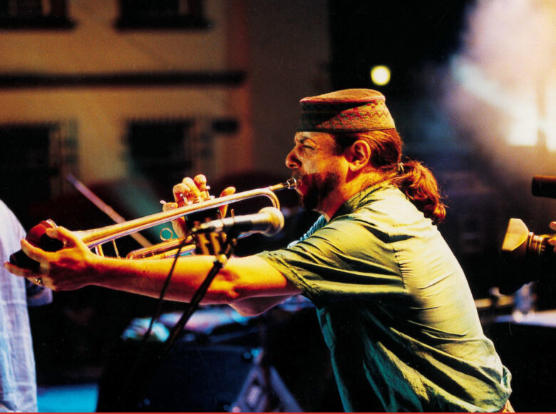 Wieczór w mieście. Na plenerowej scenie mężczyzna grający na puzonie. W tle kamienice.