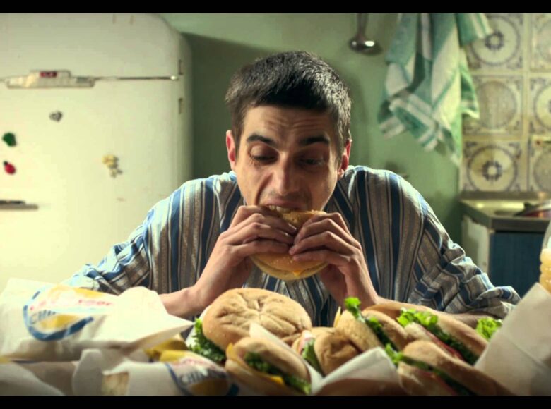 Kadr z filmu przedstawia mężczyznę w koszuli paski, który siedzi przed stołem, na którym leży stos burgerów i butelka soku pomarańczowego. Mężczyzna łapczywie je jednego z burgerów. Za nim znajduje się lodówka oraz zlew, wypełniony brudnymi naczyniami.