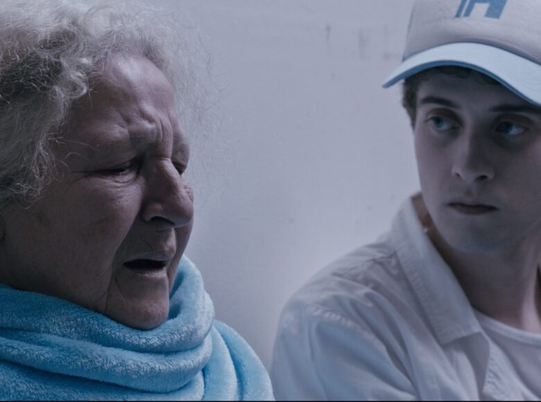 Kadr z filmu. Przy białej ścianie siedzi staruszka z owiniętym wokół szyi niebieskim szalikiem. Ma zmartwiona minę. Obok niej siedzi młody chłopak w białej koszuli i w czapce z daszkiem. Patrzy z powagą na staruszkę.