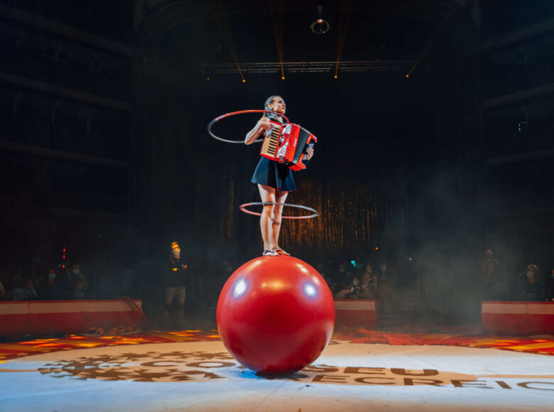 Zdjęcie przedstawia cyrkową scenę. Na dużej, czerwonej piłce stoi dziewczyna. Na jej kolanach i szyi kręcą się hula hoopy. Jednocześnie dziewczyna gra na akordeonie.