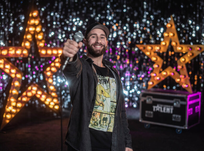 Zdjęcie przedstawia mężczyznę w rozpiętej koszuli i czapce z daszkiem. Mężczyzna uśmiecha się, jedną rękę, w której trzyma mikrofon wyciąga przed siebie. Za nim znajdują się dwie duże gwiazdy z przymocowanymi do nich żarówkami.