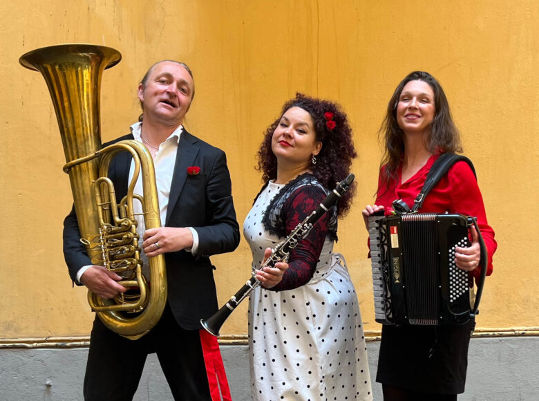 Zdjęcie przedstawia grupę muzyków: dwie kobiety i mężczyznę oraz ich instrumenty: akordeon, flet i tubę . Grupa stoi na tle żółtej ściany.