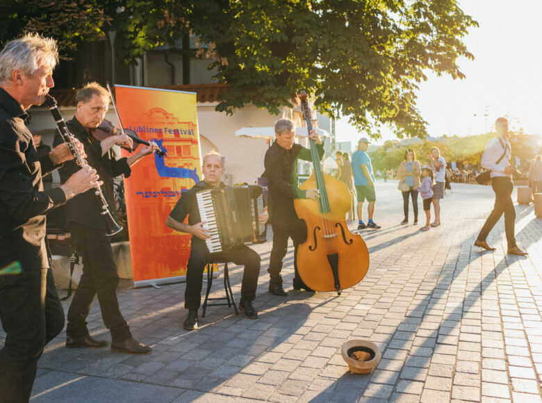 Letni dzień. Na chodniku przed zabytkowym budynkiem czterech mężczyzn w czarnych ubraniach gra na instrumentach muzycznych. Przed nimi leży kapelusz, do którego można wrzucać datki.
