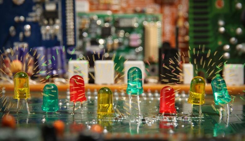 Na pierwszym planie metalowy blat. Na blacie umieszczonych kilka małych, szklanych, kolorowych przedmiocików w kształcie czopków. W tle kilka rozmazanych, kolorowych obiektów, które wyglądają jak jakieś urządzenia elektroniczne.