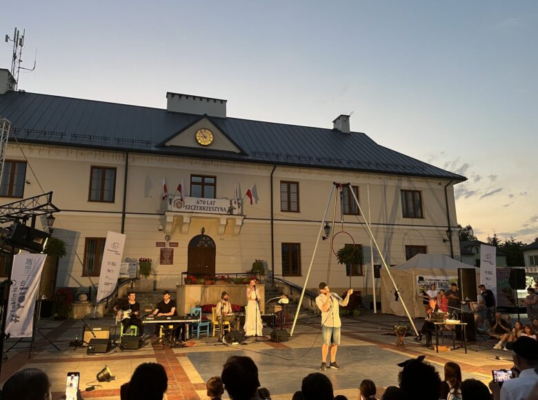 Zdjęcie przedstawia grupę ludzi zebranych wokół miejskiego placu, na którym występują muzycy i grupa cyrkowców. Na placu znajduje się budynek władz miejskich i biały namiot. Zachodzi słońce.