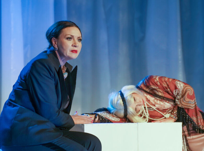 Spektakl teatralny. Dwie kobiety. Jedna, siedząca, ubrana w męski garnitur. Druga, pół-siedząca, pół-leżąca, ubrana w elegancką sukienkę i biżuterię w stylu dwudziestolecia międzywojennego.