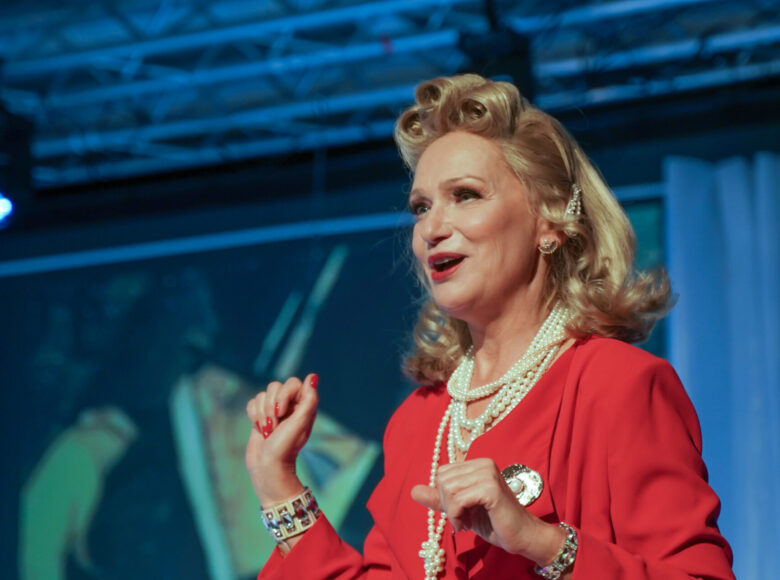 Spektakl teatralny. Zbliżenie na twarz i ramiona kobiety. Kobieta ma pomalowane na czerwono usta i paznokcie i ubrana jest w czerwony żakiet. Nosi wyrazistą, luksusową biżuterię. Ma jasne, rozpuszczone włosy z trzema lokami zakręconymi nad czołem. Za kobietą fragment scenografii w kolorach niebieskim i zielonym.