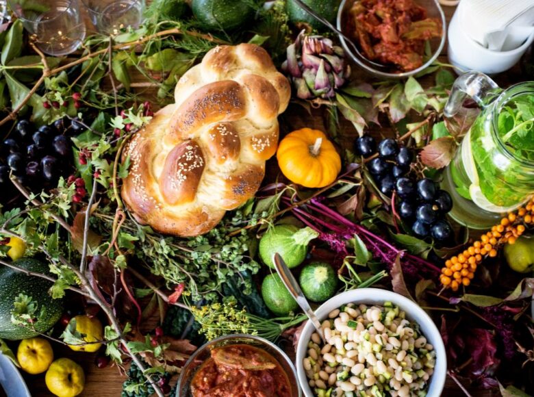 Na fotografii wykonanej z góry składniki kulinarne: chałka. dynia, okrągłe cukinie, pigwy, gałązki różnych ziół. Wśród nich stoi kilka naczyń z gotowymi potrawami.