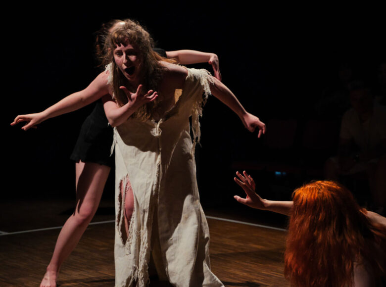 Na zdjęciu znajduje się kobieta w czerwonym kostiumie. Ma długie brązowe włosy, jej ciało jest napięte, ma ugięte kolana, przymknięte oczy, a twarz wykrzywiona jest w grymasie.