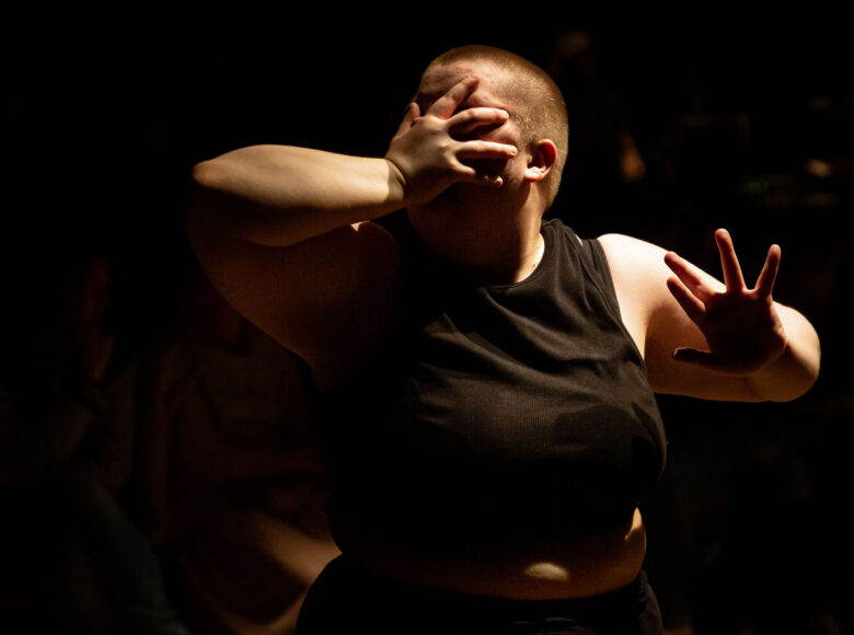 Kobieta zakrywa twarz dłonią. Ubrana jest w czarny top bez rękawów, ma bardzo krótkie włosy.