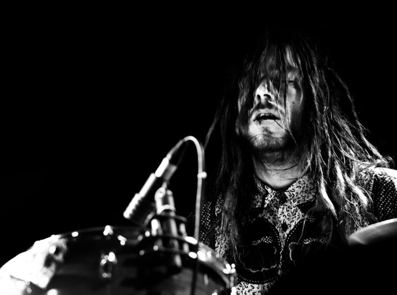 Czarno-białe zdjęcie. Na zbliżeniu twarz perkusisty. Ma zamknięte oczy i długie potargane włosy.