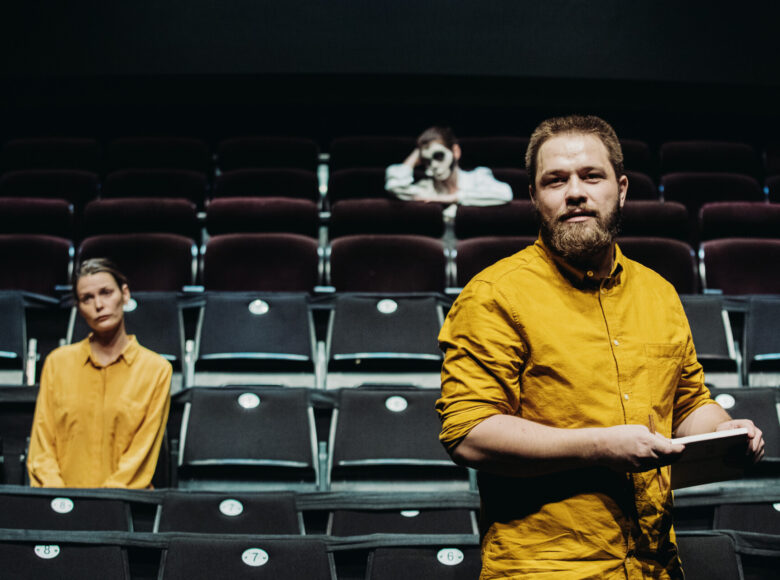 Zdjęcie ze spektaklu. Przedstawia aktorów w pomarańczowych koszulach, którzy stoją między rzędami pustej widowni.