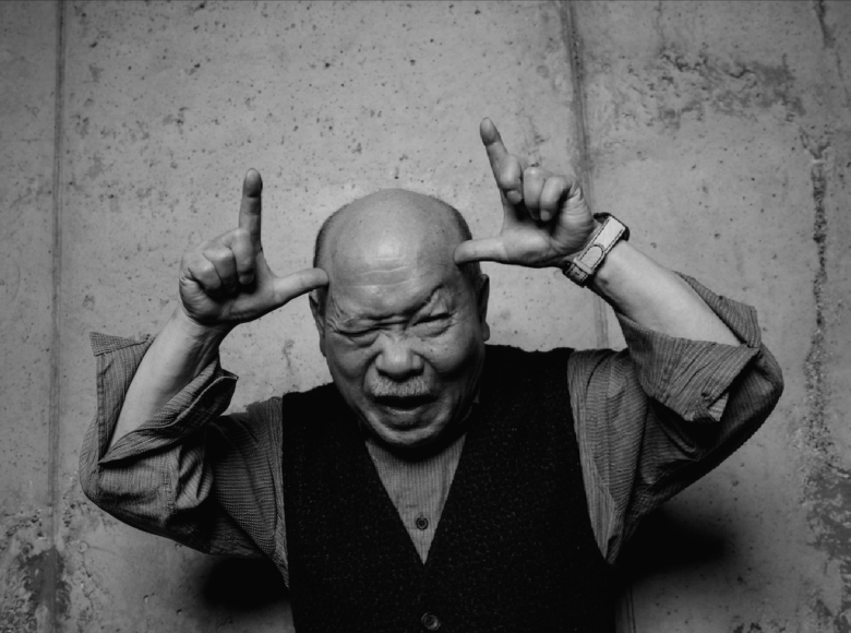 Czarno-białe zdjęcie. Przedstawia starszego mężczyznę, Azjatę. mężczyzna jest ubrany w koszulę i wełnianą kamizelkę. Mężczyzna podnosi ręce do głowy i wystawia rogi.