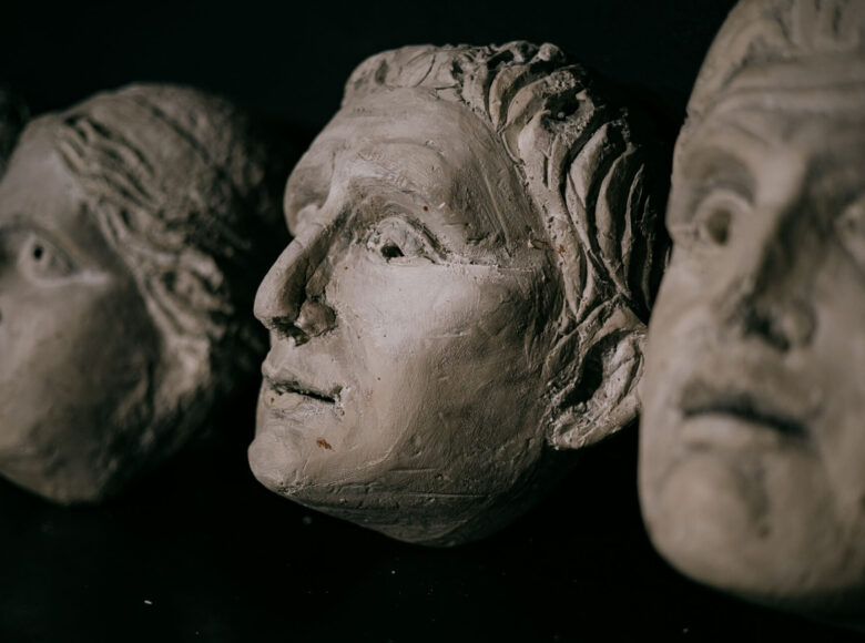 Zdjęcie przedstawia szare maski wykonane w stylu antycznym. Maski leżą ustawione w rzędzie. Tło jest czarne, jednolite.