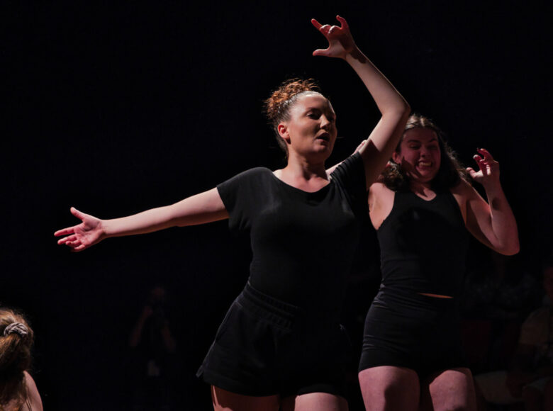 Na scenie na czarnym tle dwie biegnące tancerki tańca współczesnego. Tancerki ubrane są w czarne szorty i czarne koszulki.