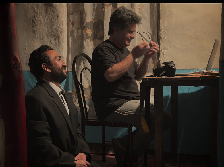 Dwóch mężczyzn w pomieszczeniu, w półmroku. Jeden siedzi na krześle, przed nim na stole stoi otwarty komputer i aparat fotograficzny. Drugi siedzi poniżej. Obaj lekko się uśmiechają.