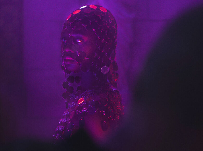 Osoba widziana od pasa w górę. Na głowie ma rodzaj nakrycia zrobionego z okrągłych, metalicznych elementów, opadających na ramiona i tors. Scena oświetlona na fioletowo.