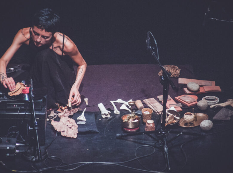 Młoda kobieta kucająca na czarnej podłodze. Przed sobą ma rozłożony gramofon, mikrofony, statywy oraz kości różniej wielości, misy mosiężne, kawałki drewna.