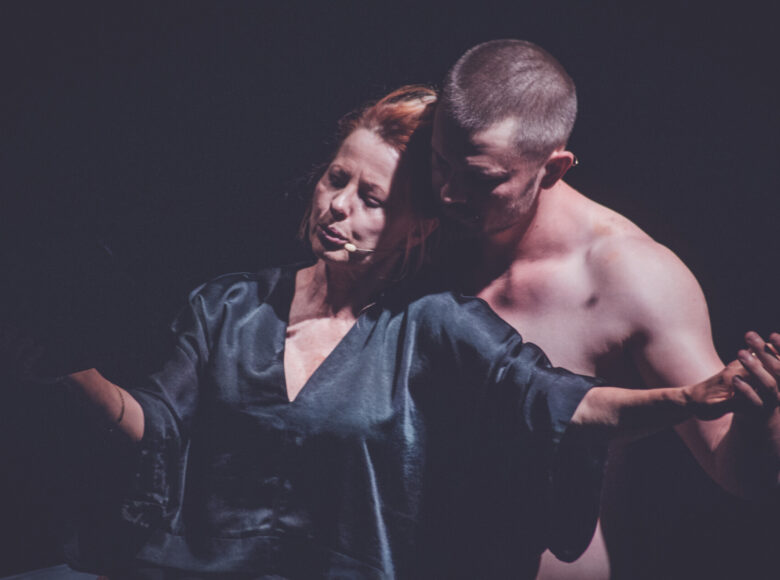 Na zdjęciu kobieta ubrana w czarną, prostą suknię oraz nagi mężczyzna, który stoi za nią. Ręce trzymają w geście przypominającym taniec.