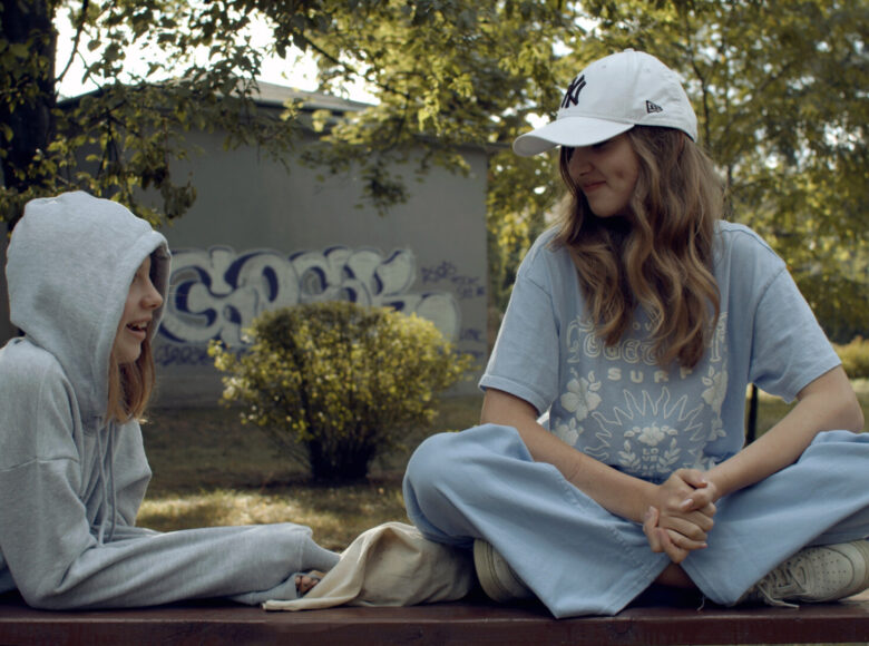 Kadr z filmu przedstawia dwie nastolatki na ławce podczas rozmowy - jedna siedzi po turecku, a druga leży podpierając się łokciami, w kapturze na głowie. W tle park, zielony plener, budynek z graffiti na ścianie.