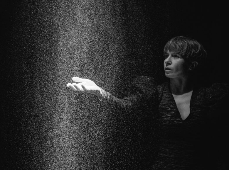 Czarno-białe zdjęcie. Kobieta z wyciągniętą przed siebie dłonią. Na jej dłoń pada snop światła, w którym unoszą się liczne drobinki kurzu.