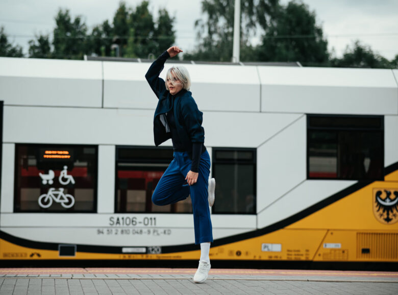 Zdjęcie przedstawia tancerkę, która znajduje się na Dworcu Kolejowym. Za nią na torach stoi pociąg.