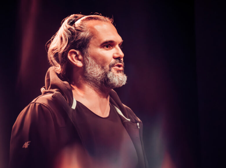 Zdjęcie przedstawia mężczyznę, który stoi bokiem i coś mówi. Jet ubrany w ciemna bluzę.