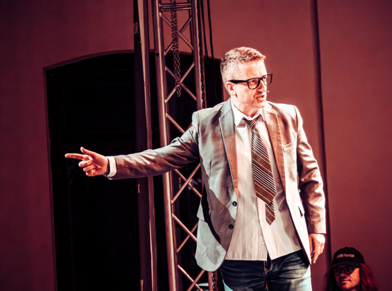 Zdjęcie przedstawia mężczyznę okularach, marynarce i krawacie. Mężczyzna stoi na scenie i wskazuje na cos ręką.