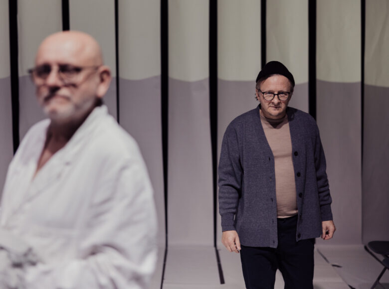 Scena teatralna. Na pierwszym planie mężczyzna w fartuchu lekarskim. Za nim stoi mężczyzna w swetrze, ciemnych spodniach, czapce na głowie i okularach.