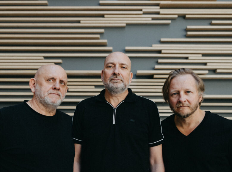 Na zdjęciu trzech dojrzałych mężczyzn w czarnych koszulkach na tle szarej ściany, ozdobionej kompozycję geometryczną z listewek.