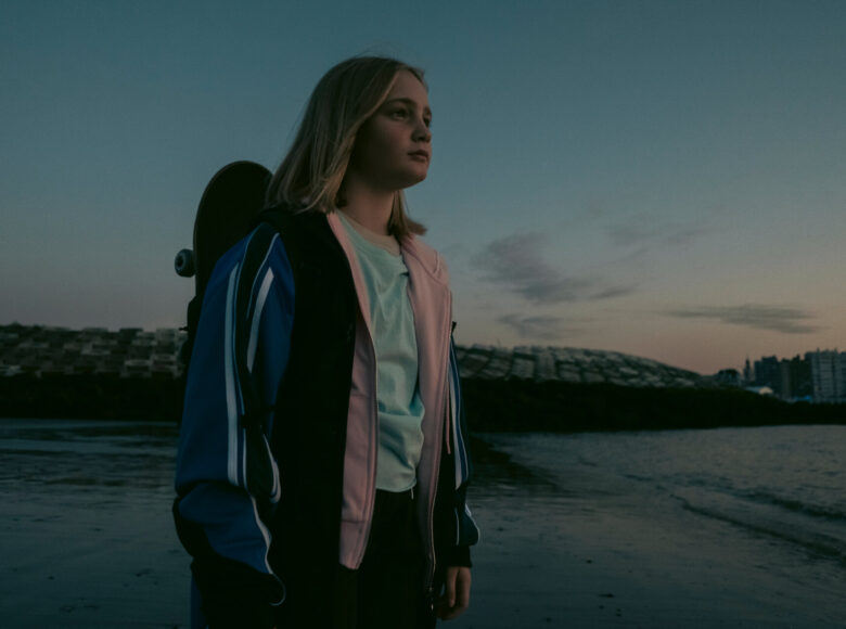 Kadr z filmu przedstawia Lenę, główną bohaterkę filmu stojącą na plaży nad brzegiem morza, w luźnym ubraniu i z deskorolką w plecaku wpatrującą się w dal.