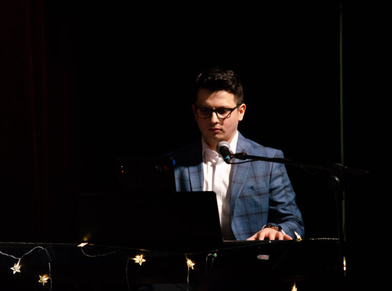 Zdjęcie przedstawia młodego, elegancko ubranego mężczyznę, który gra na pianinie. Tło ciemne, jednolite.