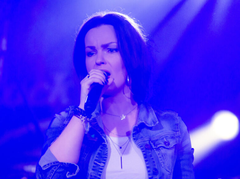 Zdjęcie przedstawia kobietę, która znajduje się na scenie. W ręce trzyma mikrofon i śpiewa. Na scenę pada niebieskie światło.