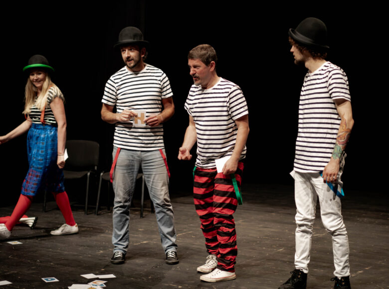 Na scenie stoi trzech mężczyzn ubranych w jednakowe koszulki w czarno-białe paski. Obok nich idzie kobieta w czarnym kapeluszu na głowie.