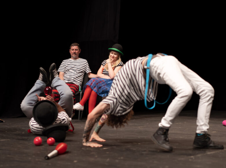 Na scenie znajduje się grupa ludzi w koszulkach w biało-czarne paski. Dwie osoby siedzą na krzesłach, dwie pozostałe wykonują akrobacje.
