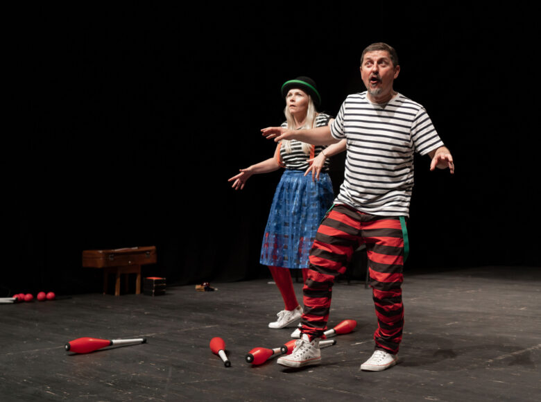 Zdjęcie przedstawia kobietę i mężczyznę. Znajdują się na scenie, oboje są ubrani w koszuli w czarno-białe paski. Na podłodze leżą czerwone maczugi do żonglerki.