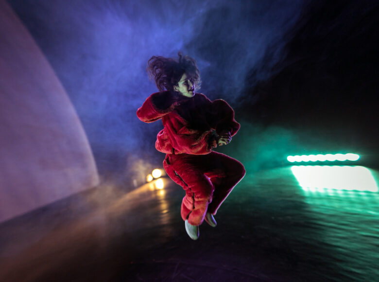 Na zdjęciu młoda dziewczyna w ciemnopomarańczowym stroju misia. Osoba w podskoku, za nią zielone i fioletowe plamy światła.