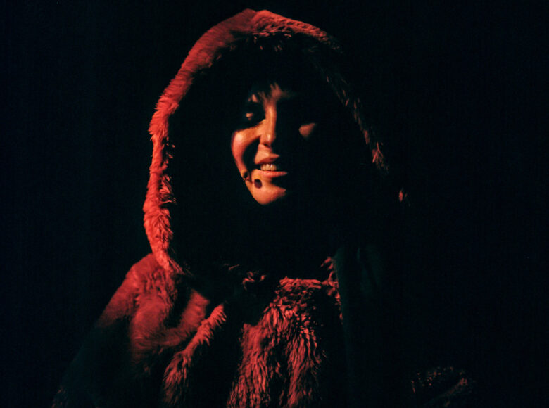 Zbliżenie na twarz dziewczyny, na głowie ma ciemnopomarańczowy kaptur. Lekko się uśmiecha.