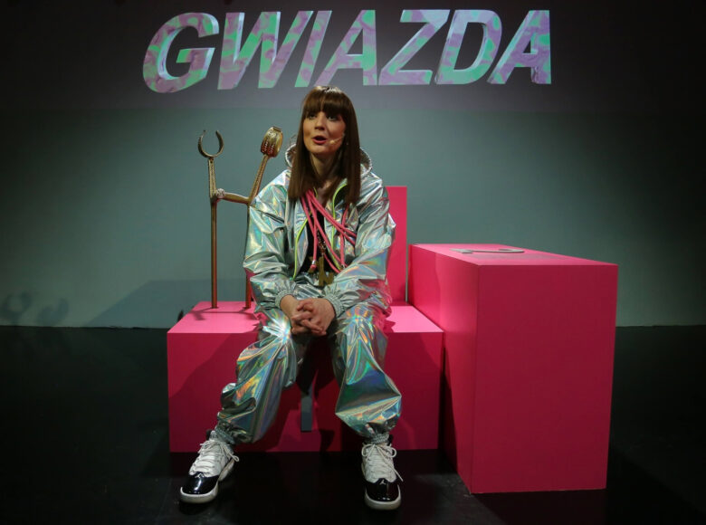 Młoda kobieta ubrana w błyszczący dres, siedzi na różowych kubikach. W tle za nią napis „Gwiazda”.