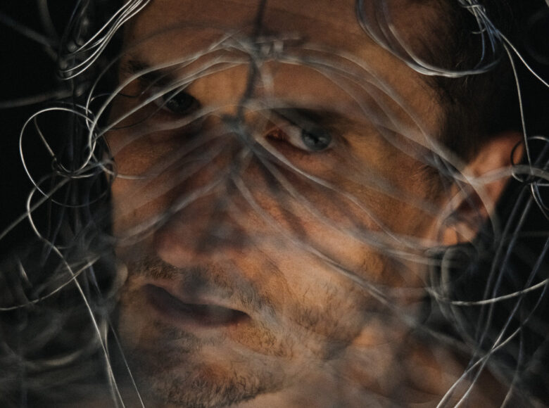 Zdjęcie przedstawia twarz mężczyzny na zbliżeniu. Mężczyzna patrzy przed siebie. Twarz przysłaniają metalowe druty.