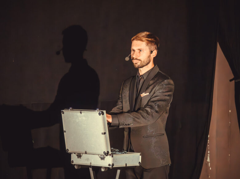 Zdjęcie przedstawia mężczyznę w ciemnym, błyszczącym garniturze. Mężczyzna wyciąga coś ze srebrnej, otwartej walizki. Na ścianę pada jego cień.