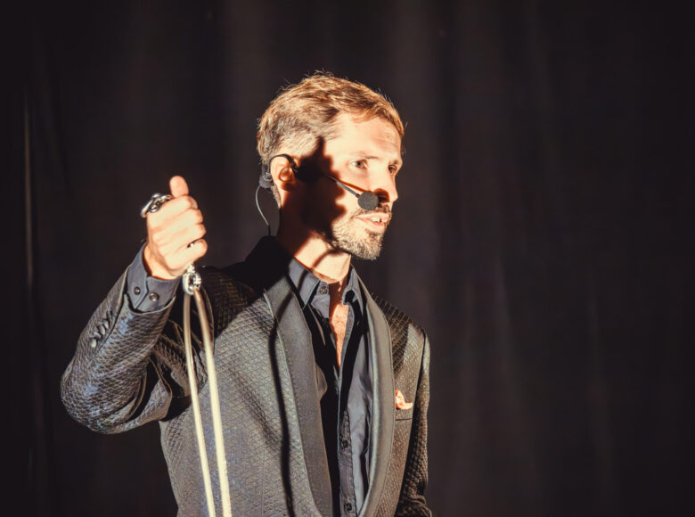 Na scenie stoi mezczyna w czarnym, błyszczącym garniturze. Pada na niego jasne światło. W ręce trzyma metalowy, srebrny łańcuch.