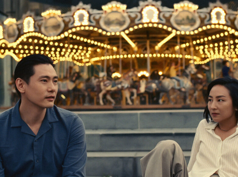 Na zdjęciu kobieta i mężczyzna o azjatyckich rysach twarzy. W tle za nimi staromodna karuzela oświetlona sznurami żarówek.