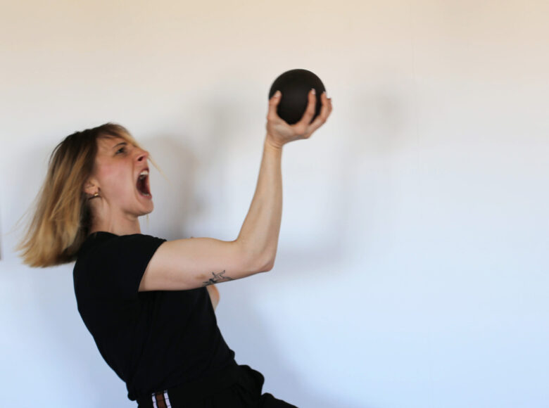 Zdjecie przedstawia młodą kobietę w czarnym sportowym ubraniu. Kobieta trzyma w jednej ręce czarną kulę. Ma szeroko otwarte usta jakby krzyczała.