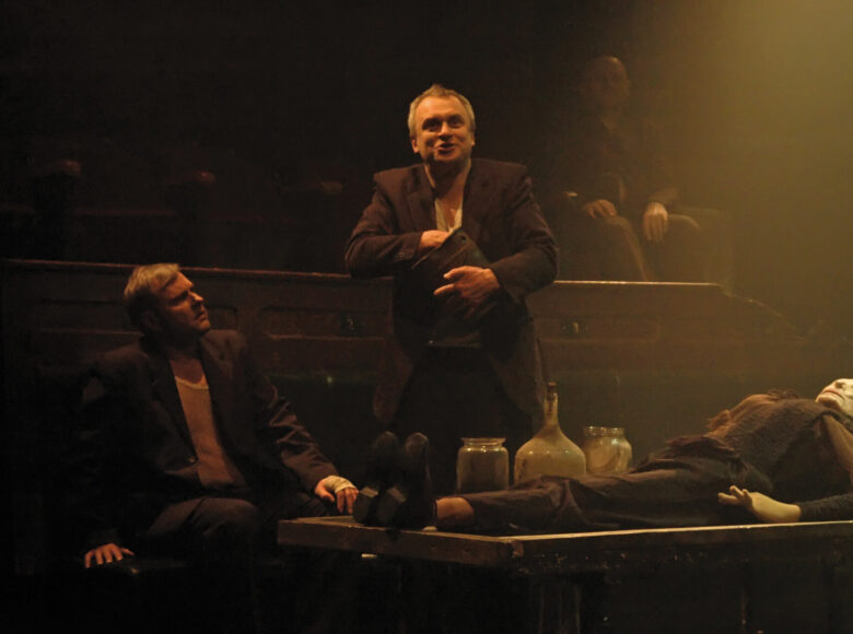 Dwóch mężczyzn przy stole na którym leży kukła. Jeden siedzi, drugi stojąc mówi, trzyma w rękach garnek, z którego coś wyjmuje.