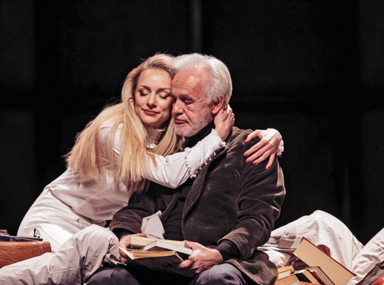 Scena ze spektaklu. Młoda kobieta, blondynka, obejmuje za szyję starszego, siwego mężczyznę, który siedzi obok niej. Wokół nich rozrzucone książki.