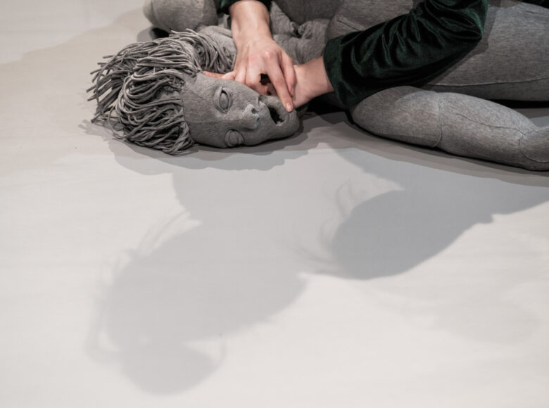 Na zbliżeniu twarz szarej kukły. Dłonie niewidocznej w kadrze osoby przyciskają twarz do szarej podłogi.